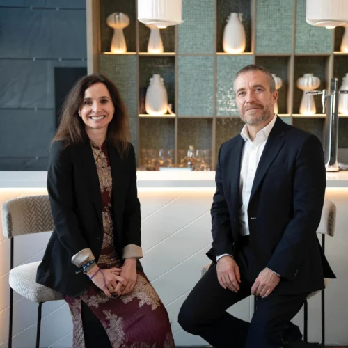 Miguel Sanz, director general de Turespaña, y Teresa Zamora, directora de Marketing y Comunicación de ILUNION Hotels, posan sentados frente a una barra de bar elegante con estanterías iluminadas que exhiben jarrones blancos.