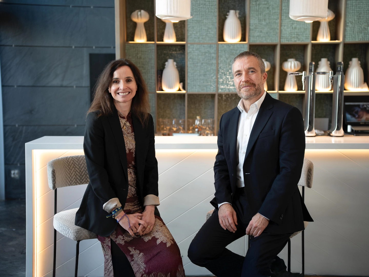 Miguel Sanz, director general de Turespaña, y Teresa Zamora, directora de Marketing y Comunicación de ILUNION Hotels, posan sentados frente a una barra de bar elegante con estanterías iluminadas que exhiben jarrones blancos.