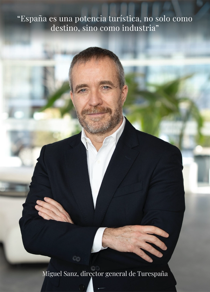 Retrato de medio cuerpo de Miguel Sanz, director general de Turespaña, posando con los brazos cruzados y vistiendo un traje oscuro. En la parte superior de la imagen se lee la cita: 'España es una potencia turística no solo como destino, sino como industria'. El fondo es un interior de oficina luminoso con plantas difuminadas.