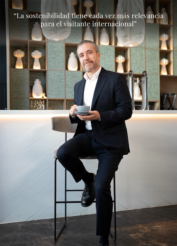 Miguel Sanz posa sentado en un taburete alto frente a una barra, sosteniendo una taza de café. Al fondo se observa una estantería con piezas de cerámica decorativa. En la parte superior, la cita: 