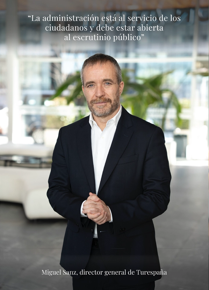Retrato de medio cuerpo de Miguel Sanz, director general de Turespaña, vistiendo un traje oscuro y camisa blanca con las manos entrelazadas. En la parte superior de la imagen aparece la cita: 'La administración está al servicio de los ciudadanos y debe estar abierta al escrutinio público'. El fondo muestra un espacio interior moderno y luminoso con plantas.