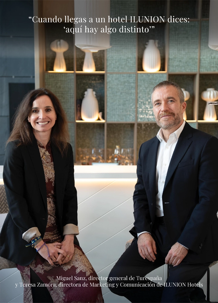Miguel Sanz, director general de Turespaña, y Teresa Zamora, directora de Marketing y Comunicación de ILUNION Hotels, posan sentados frente a una barra de bar elegante con estanterías iluminadas que exhiben jarrones blancos. En la parte superior, una cita reza: 'Cuando llegas a un hotel ILUNION dices: «aquí hay algo distinto