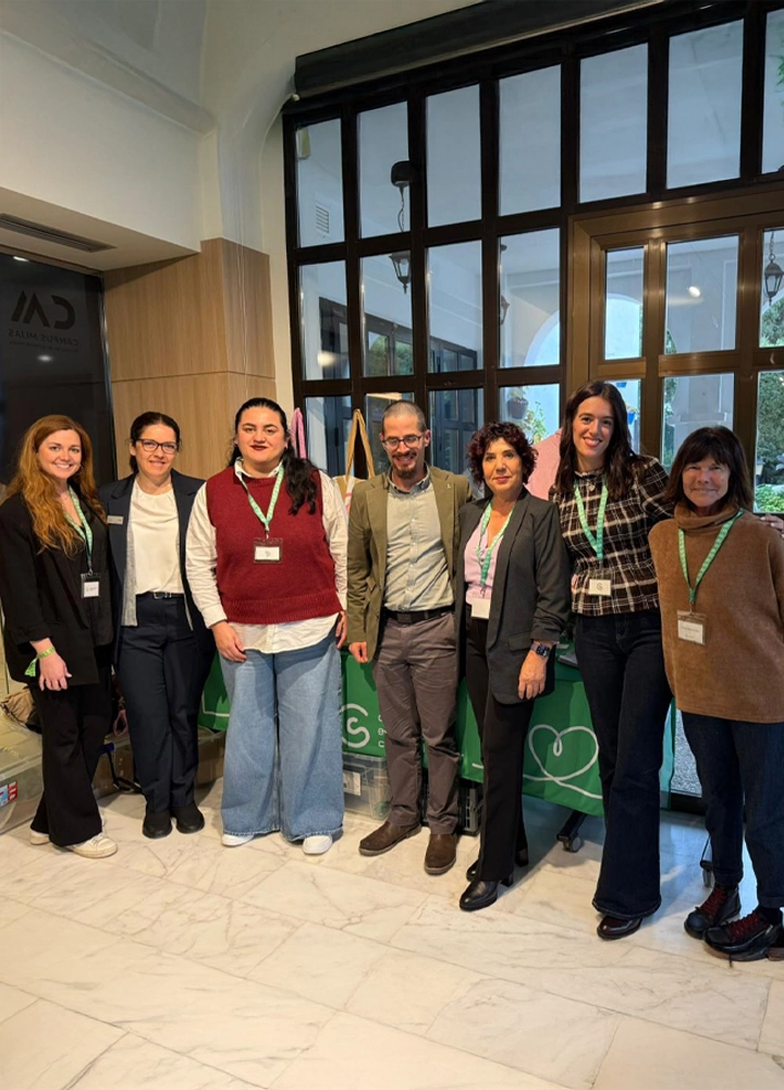 Un grupo de siete personas posa sonriente frente a un mostrador de recepción verde. Todas llevan acreditaciones con cordones verdes. El grupo está compuesto por cinco mujeres y dos hombres con vestimenta profesional variada en un vestíbulo con suelos de mármol.