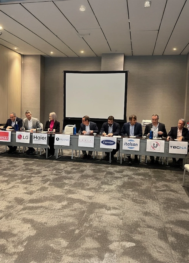 Ocho personas (siete hombres y una mujer) están sentadas en una mesa larga de conferencias frente a una pantalla de proyección blanca. Frente a cada participante hay carteles con logotipos de empresas como LG, Haier, Airzone, Carrier e Italsan. Se encuentran en una sala de reuniones con alfombra gris.
