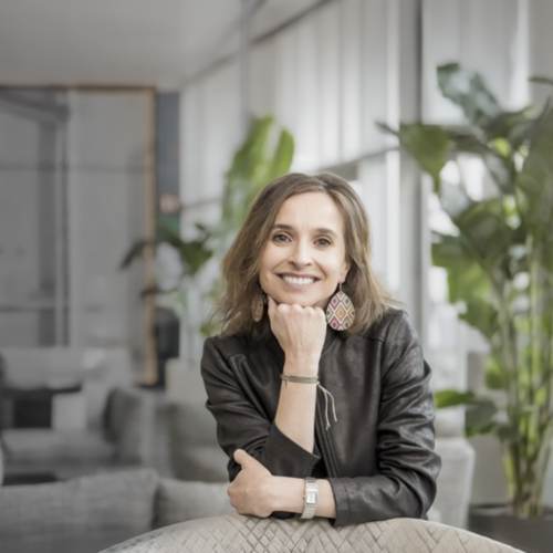 Persona con chaqueta de cuero oscura sentada en un espacio interior luminoso con plantas altas y mobiliario moderno. Apoya un brazo sobre la mesa y sostiene su barbilla con la mano, en un ambiente de oficina contemporánea y relajada.