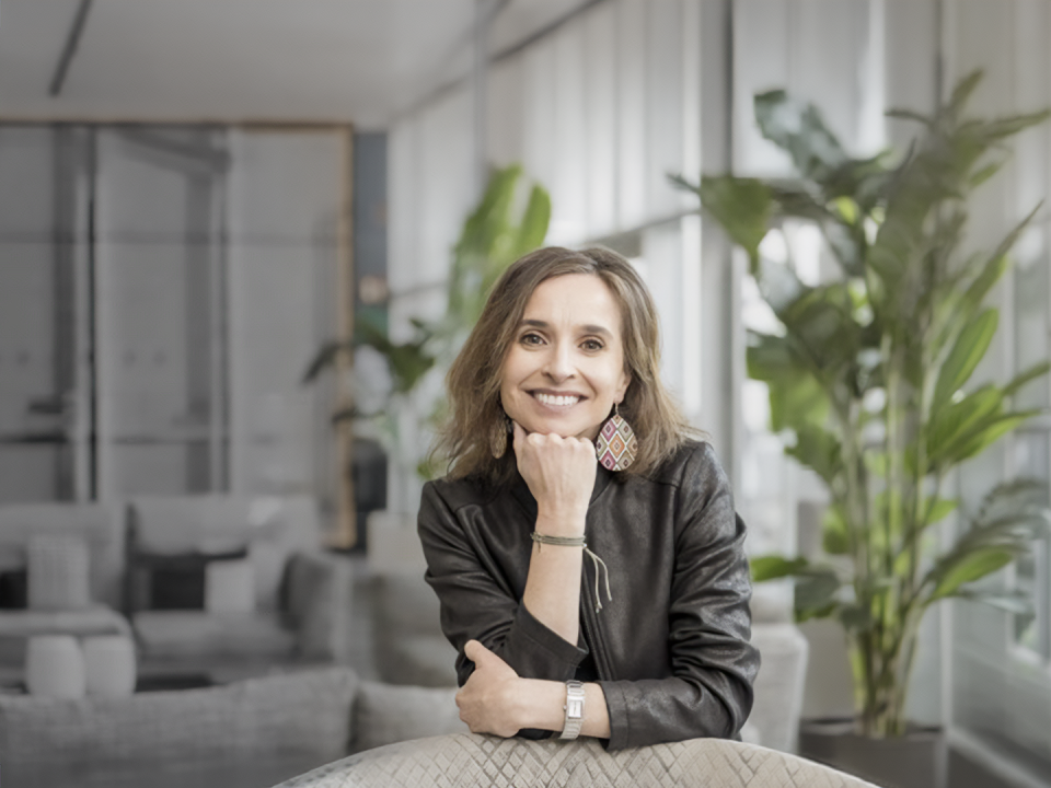 Persona con chaqueta de cuero oscura sentada en un espacio interior luminoso con plantas altas y mobiliario moderno. Apoya un brazo sobre la mesa y sostiene su barbilla con la mano, en un ambiente de oficina contemporánea y relajada.