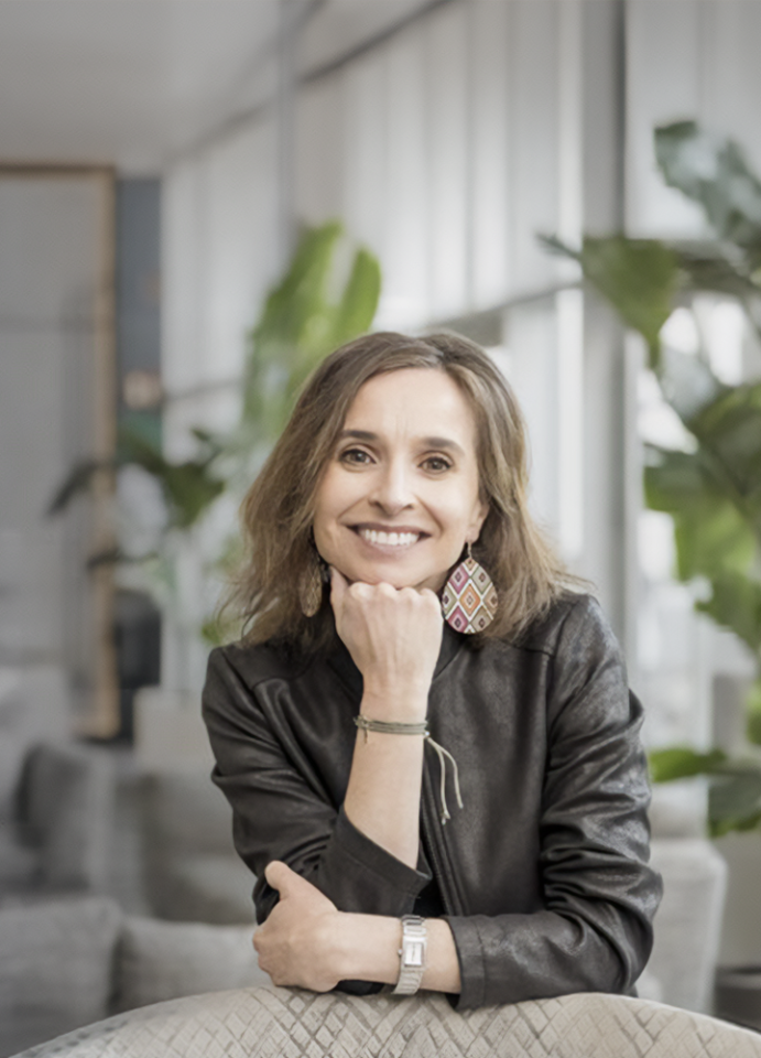 Persona con chaqueta de cuero oscura sentada en un espacio interior luminoso con plantas altas y mobiliario moderno. Apoya un brazo sobre la mesa y sostiene su barbilla con la mano, en un ambiente de oficina contemporánea y relajada.