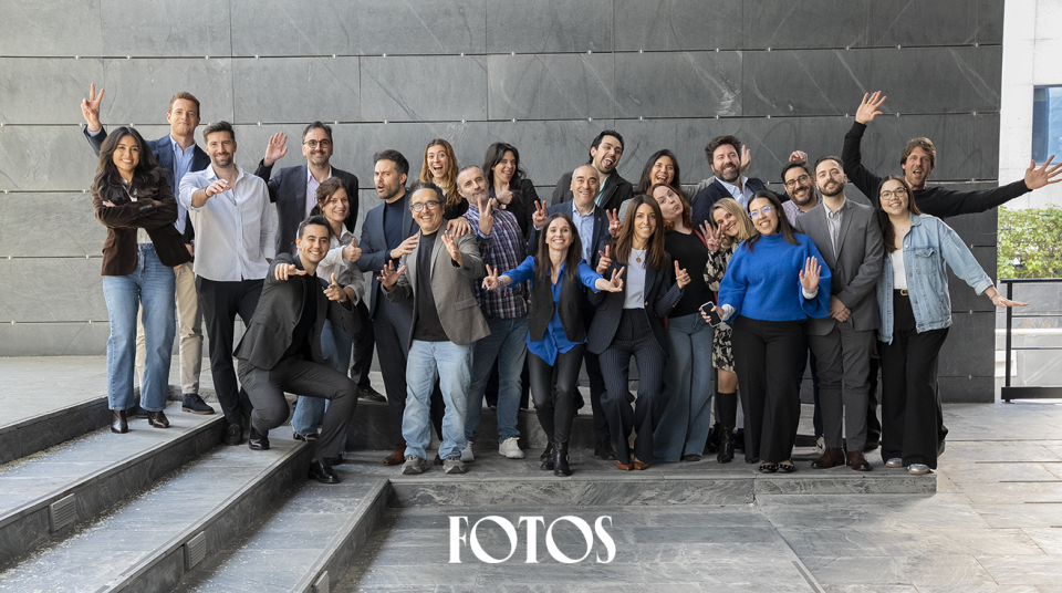 Grupo grande posando en varios niveles de escalones de piedra, todos haciendo gestos divertidos con las manos, como señales de paz o brazos alzados. El fondo es un muro gris de bloques lisos y el ambiente transmite energía y compañerismo. En la parte inferior aparece la palabra FOTOS.
