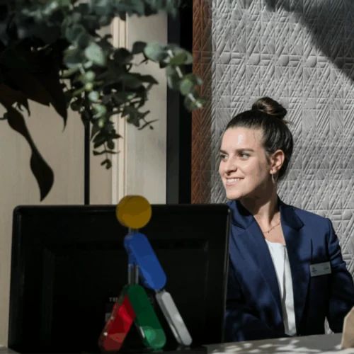 Persona vestida con blazer azul y camiseta clara, sentada tras un mostrador con un ordenador, en un interior iluminado por luz lateral. Sobre el monitor hay una pequeña figura decorativa multicolor y, al fondo, una pared con textura geométrica y plantas en la parte superior izquierda.