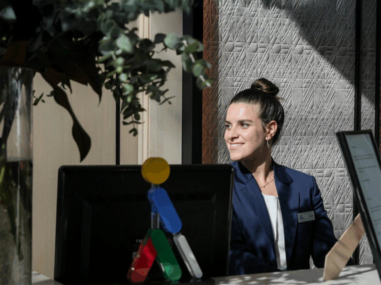 Persona vestida con blazer azul y camiseta clara, sentada tras un mostrador con un ordenador, en un interior iluminado por luz lateral. Sobre el monitor hay una pequeña figura decorativa multicolor y, al fondo, una pared con textura geométrica y plantas en la parte superior izquierda.