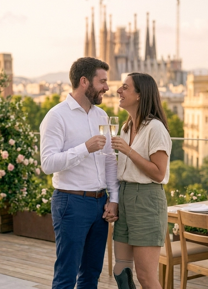 Una pareja joven brindando con copas de champán en una terraza de madera al atardecer, con la icónica Basílica de la Sagrada Familia de Barcelona de fondo. El hombre, con barba y camisa blanca, y la mujer, con una blusa beige y pantalones cortos verdes, se miran y ríen mientras se toman de la mano. La mujer tiene una prótesis en su pierna derecha. El entorno incluye plantas con flores rosas y mesas de comedor, todo bajo una luz cálida y suave.