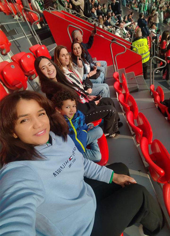 Varias personas sentadas en filas de asientos rojos dentro de un estadio, vistas en diagonal desde arriba. Visten ropa informal y observan un evento deportivo, con público y personal de seguridad visibles en las gradas del fondo. Ambiente animado y de asistencia a un partido.