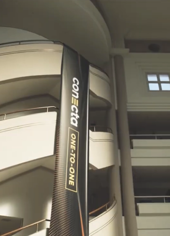 Gran atrio interior con varios niveles de pasarelas curvas y barandillas claras. Desde uno de los pisos cuelga una lona vertical negra con el texto “conecta” en blanco y “ONE-TO-ONE” en un recuadro amarillo. La luz natural entra desde la parte superior del edificio.