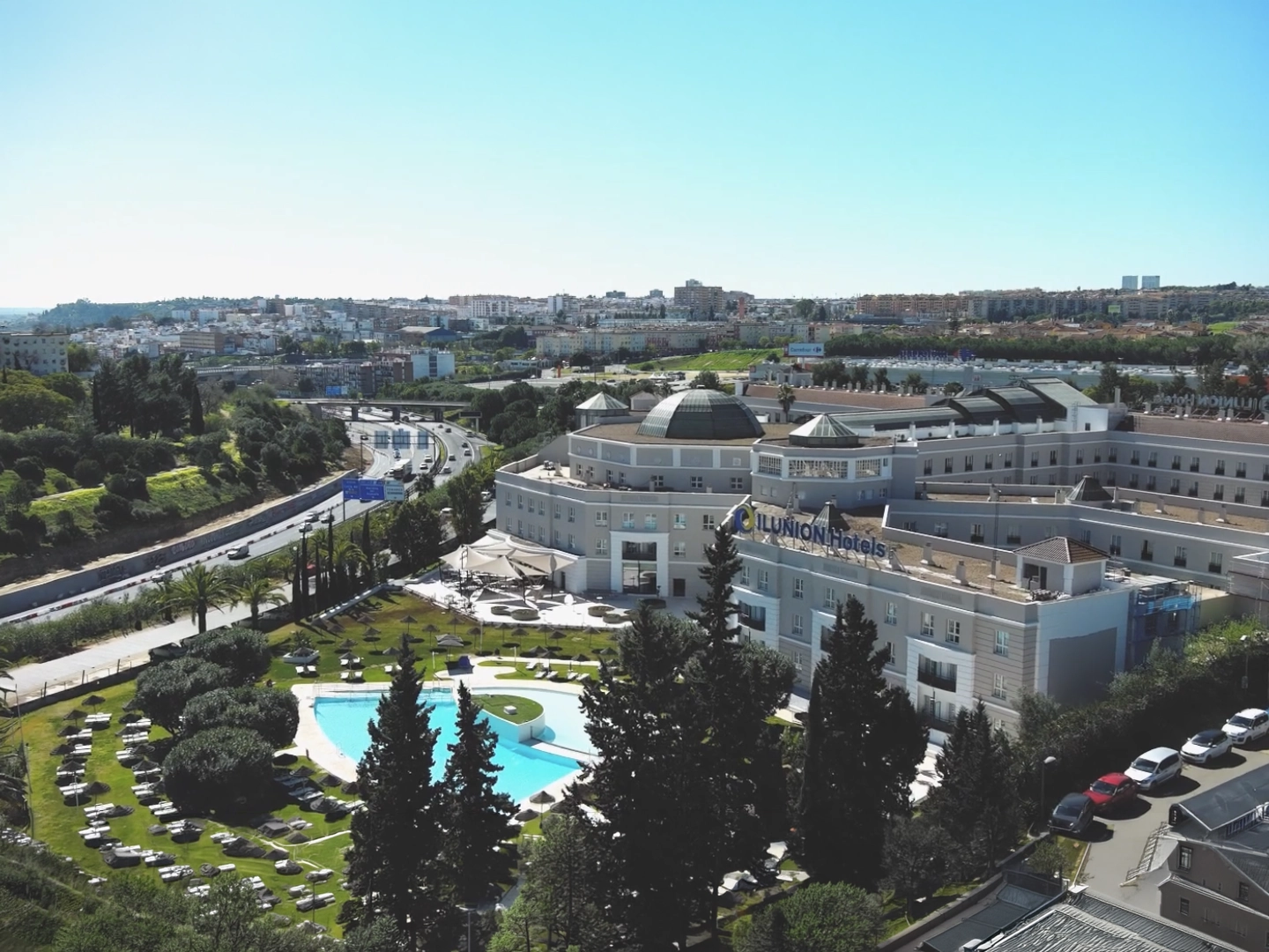 Vista aérea de un gran complejo hotelero rodeado de jardines cuidados, con una piscina de forma orgánica y varias zonas de descanso al aire libre. El edificio principal, de varios niveles y tonos claros, se sitúa junto a una carretera que serpentea entre áreas verdes. Al fondo se extiende una ciudad con edificios residenciales bajo un cielo despejado.