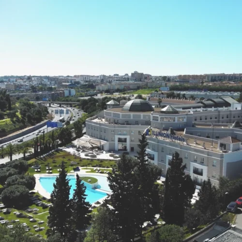 Vista aérea de un gran complejo hotelero rodeado de jardines cuidados, con una piscina de forma orgánica y varias zonas de descanso al aire libre. El edificio principal, de varios niveles y tonos claros, se sitúa junto a una carretera que serpentea entre áreas verdes. Al fondo se extiende una ciudad con edificios residenciales bajo un cielo despejado.