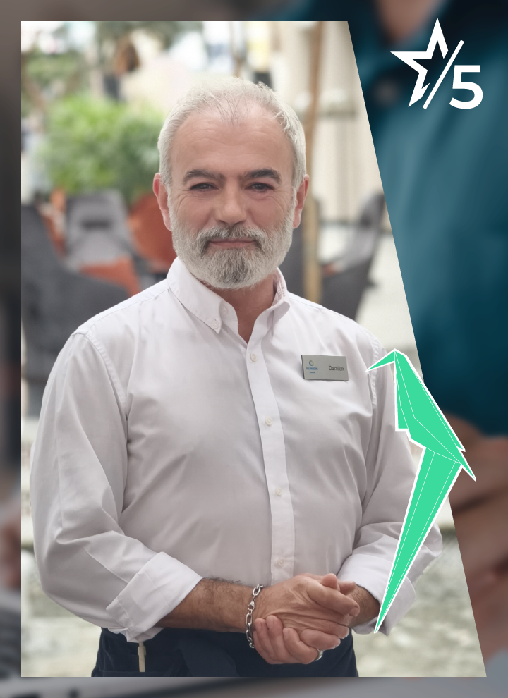 Persona de pie con camisa blanca y placa identificativa, las manos entrelazadas al frente. Fondo desenfocado con mesas y sillas de exterior, y un origami y el número 5 en la esquina superior derecha.