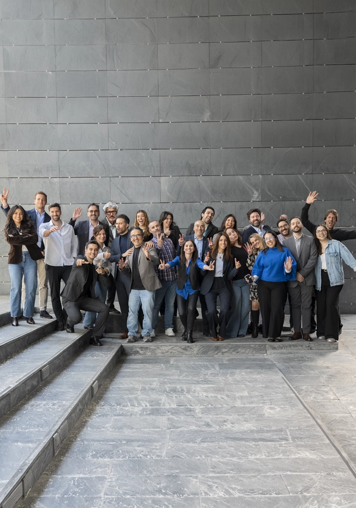 Grupo grande posando en varios niveles de escalones de piedra, todos haciendo gestos divertidos con las manos, como señales de paz o brazos alzados. El fondo es un muro gris de bloques lisos y el ambiente transmite energía y compañerismo.