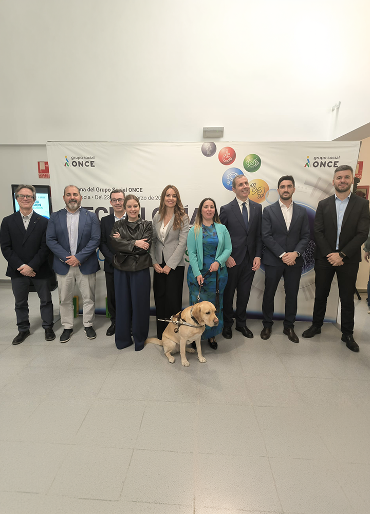 Grupo de personas adultas posando de pie en un espacio interior, delante de un panel con el logotipo de Grupo Social ONCE y elementos gráficos de colores. Visten ropa formal y semiformal, como trajes y chaquetas. En el centro del grupo, un perro guía de color claro está sentado en el suelo, con arnés, junto a una persona. El ambiente es institucional y de acto oficial.