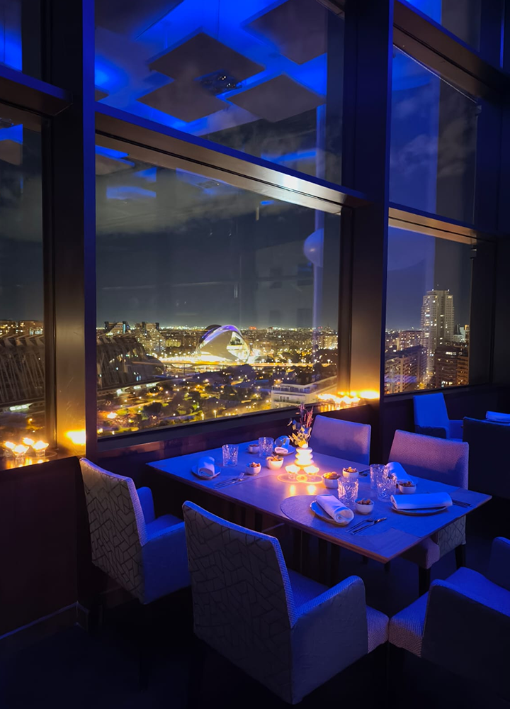 Mesa para varias personas en un restaurante elegante, junto a grandes ventanales con vistas nocturnas a una ciudad iluminada. La mesa está preparada con mantelería, copas, platos y velas encendidas, creando un ambiente íntimo. Predominan luces azules y tonos cálidos, con sillones tapizados y un entorno moderno y sofisticado.