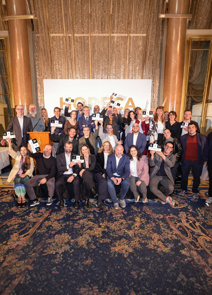 Fotografía grupal de un numeroso grupo de personas celebrando en el escenario de un salón elegante. La mayoría de los asistentes están de pie o sentados en la parte frontal, muchos de ellos alzando trofeos rectangulares con el logo 