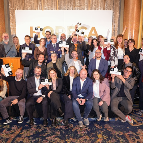 Fotografía grupal de un numeroso grupo de personas celebrando en el escenario de un salón elegante. La mayoría de los asistentes están de pie o sentados en la parte frontal, muchos de ellos alzando trofeos rectangulares con el logo 