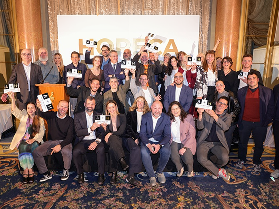 Fotografía grupal de un numeroso grupo de personas celebrando en el escenario de un salón elegante. La mayoría de los asistentes están de pie o sentados en la parte frontal, muchos de ellos alzando trofeos rectangulares con el logo "A + H". De fondo, hay un panel blanco con la palabra "HORECA" en letras doradas y cortinajes brillantes que cubren la pared. La sala tiene un estilo clásico con alfombras de patrones florales y columnas de mármol.