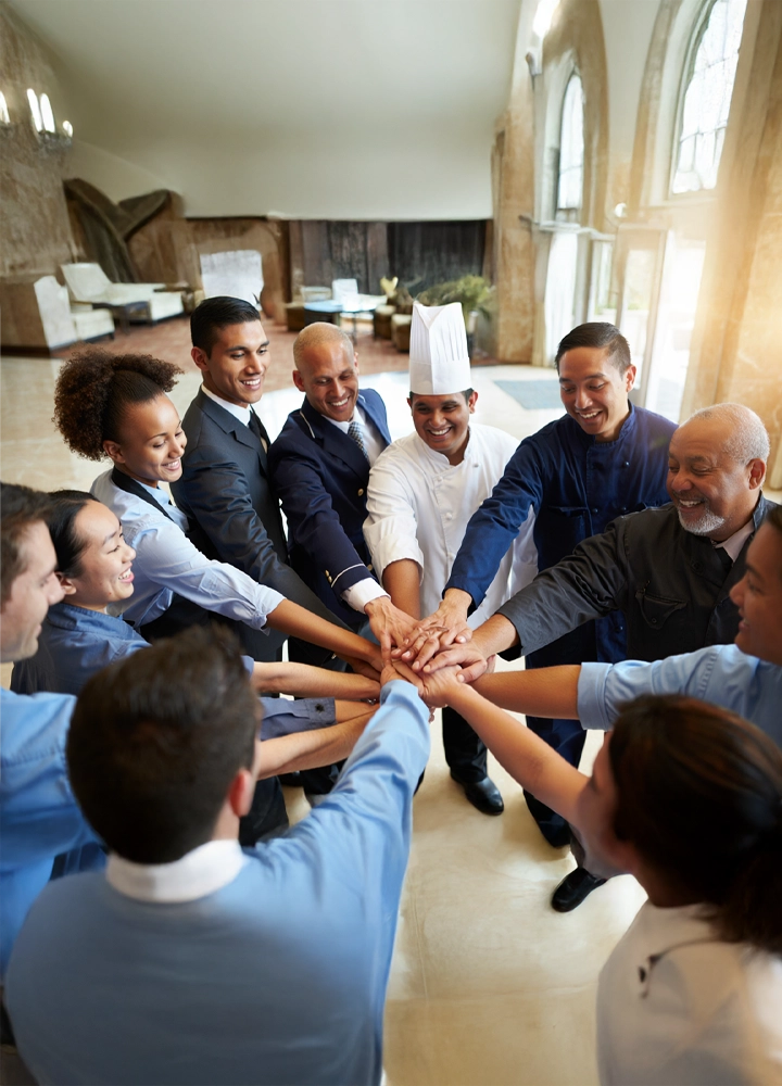 Un grupo diverso de empleados de un hotel, incluyendo personal de cocina, recepción, limpieza y mantenimiento, están reunidos en un círculo en el vestíbulo, uniendo sus manos en el centro en un gesto de unidad y trabajo en equipo. Todos sonríen en un ambiente luminoso y colaborativo.