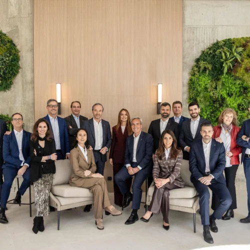 Grupo numeroso de personas posando en un espacio interior elegante, con dos asientos frontales ocupados y el resto de pie alrededor. Visten ropa formal y están situados frente a una pared clara decorada con paneles verticales y dos grandes círculos de vegetación.
