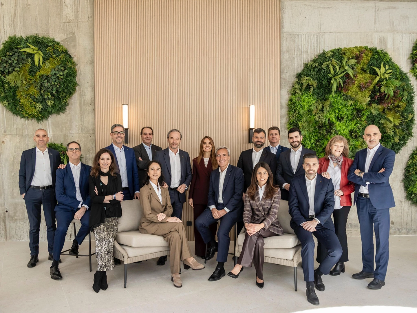 Grupo numeroso de personas posando en un espacio interior elegante, con dos asientos frontales ocupados y el resto de pie alrededor. Visten ropa formal y están situados frente a una pared clara decorada con paneles verticales y dos grandes círculos de vegetación.