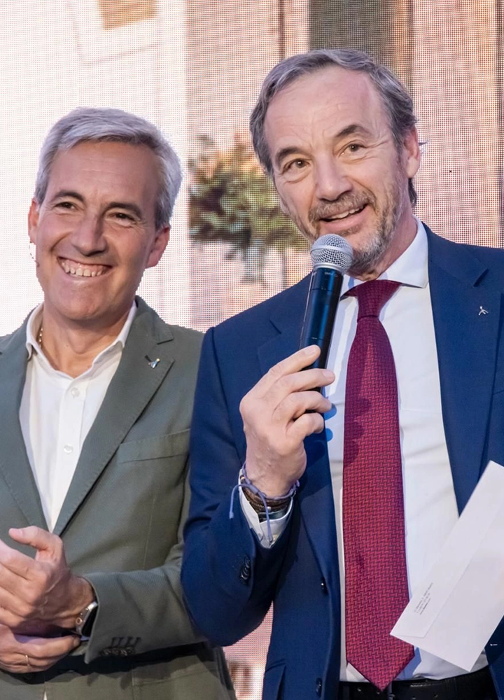 Fotografía de dos directivos sonrientes en un escenario. Un hombre de traje azul marino habla por un micrófono, mientras el otro, con una chaqueta verde oliva, le acompaña y sonríe.