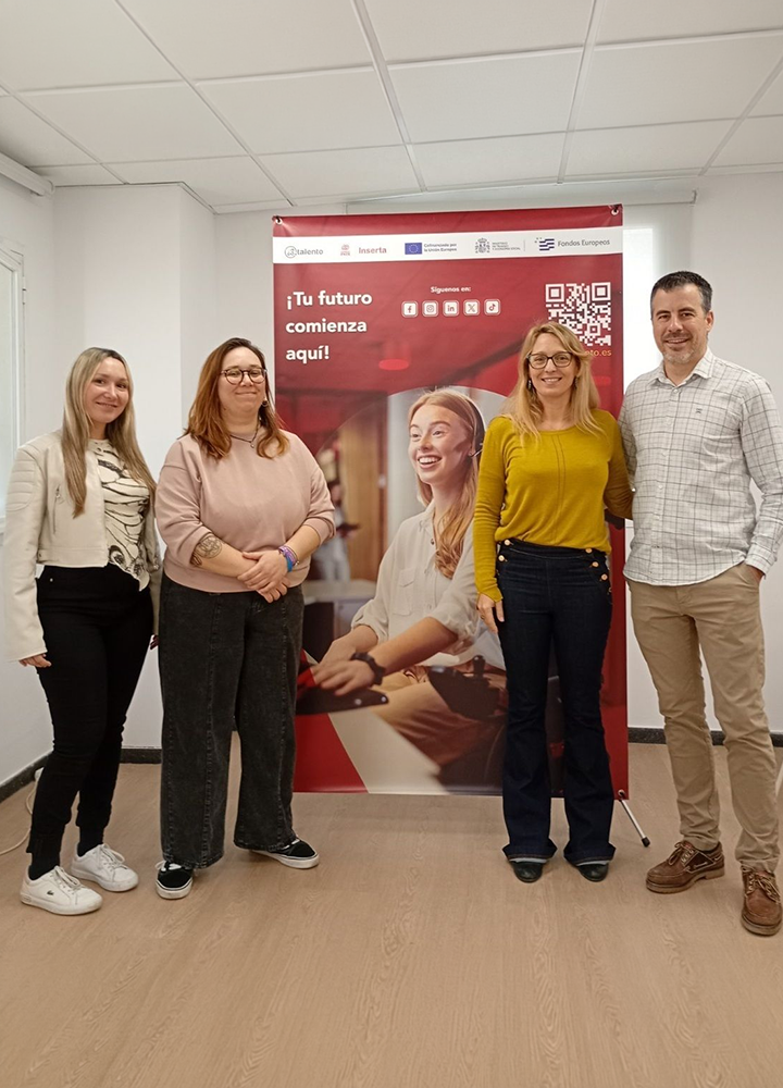 Cinco personas adultas de pie en una sala interior con suelo de madera y techo blanco. Posan frente a un roll‑up rojo con el texto “¡Tu futuro comienza aquí!”, logotipos institucionales en la parte superior y un código QR a la derecha. Visten ropa informal y semiformal, con chaquetas, jerséis y pantalones. El ambiente es profesional y formativo, propio de una presentación o actividad de orientación.