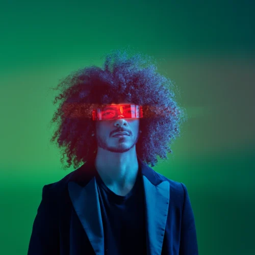 Retrato de estudio de un hombre con peinado afro voluminoso y vello facial, vistiendo una chaqueta de traje oscura. Lleva unas gafas tecnológicas rectangulares de color rojo translúcido que emiten un brillo intenso sobre sus ojos. La iluminación de la escena es dramática, con tonos azules y verdes neón de estilo futurista.