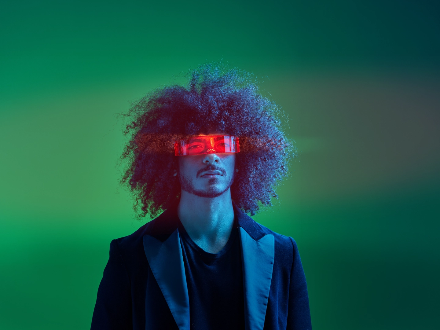 Retrato de estudio de un hombre con peinado afro voluminoso y vello facial, vistiendo una chaqueta de traje oscura. Lleva unas gafas tecnológicas rectangulares de color rojo translúcido que emiten un brillo intenso sobre sus ojos. La iluminación de la escena es dramática, con tonos azules y verdes neón de estilo futurista.