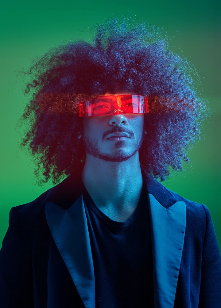 Retrato de estudio de un hombre con peinado afro voluminoso y vello facial, vistiendo una chaqueta de traje oscura. Lleva unas gafas tecnológicas rectangulares de color rojo translúcido que emiten un brillo intenso sobre sus ojos. La iluminación de la escena es dramática, con tonos azules y verdes neón de estilo futurista.