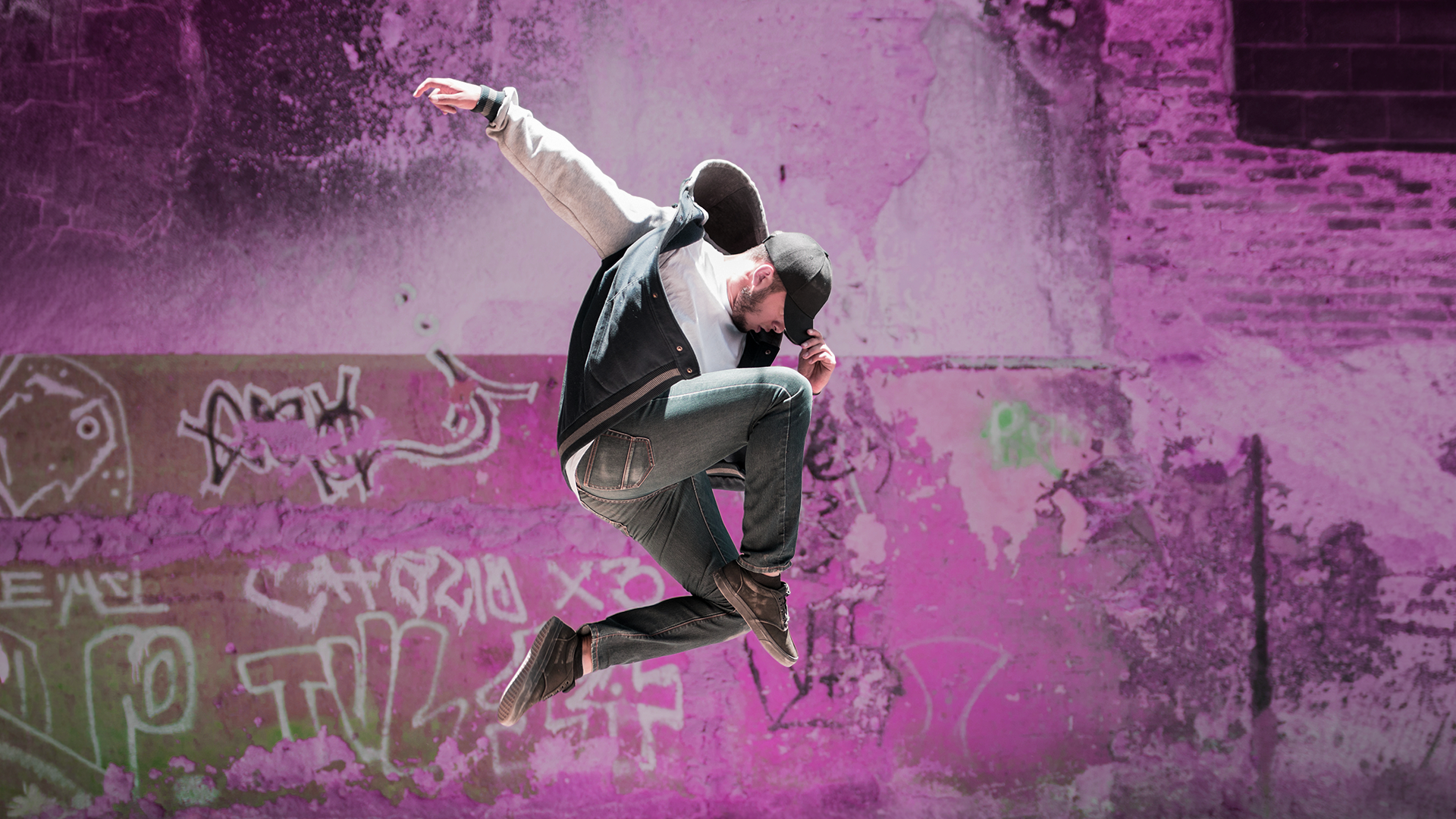 Persona saltando en el aire con una postura dinámica, brazo extendido y piernas recogidas. Viste gorra, chaqueta bicolor y vaqueros. El fondo es una pared urbana desgastada con grafitis en tonos rosas y morados.
