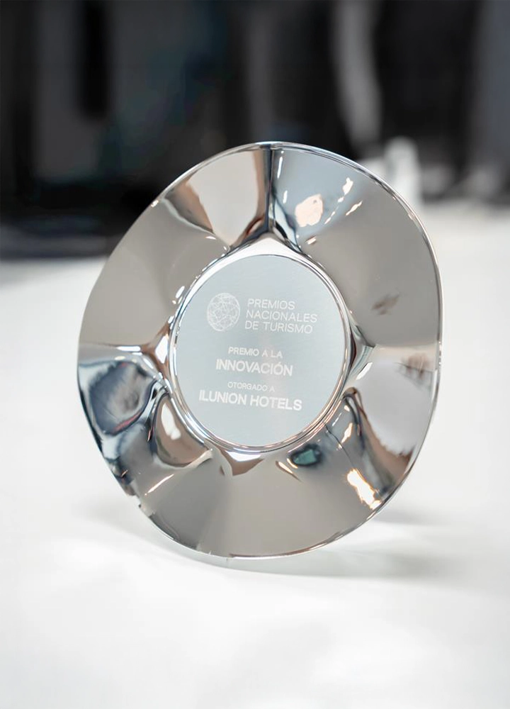 Trofeo metálico circular y brillante de los 'Premios Nacionales de Turismo'. La inscripción central indica: 'Premio a la Innovación otorgado a Ilunion Hotels'. El premio reposa sobre una superficie clara con un fondo desenfocado.