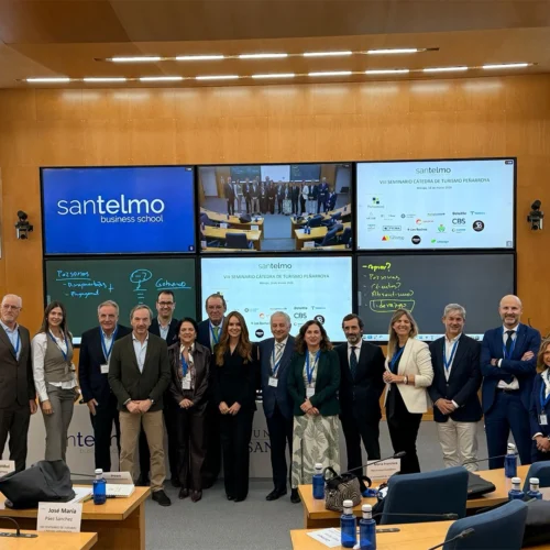 Grupo de personas posando en el centro de un aula tipo auditorio, con varias pantallas al fondo que muestran el nombre “Santelmo Business School”, logotipos de empresas colaboradoras y una imagen grupal. Mesas con tarjetas identificativas y botellas azules rodean la escena.