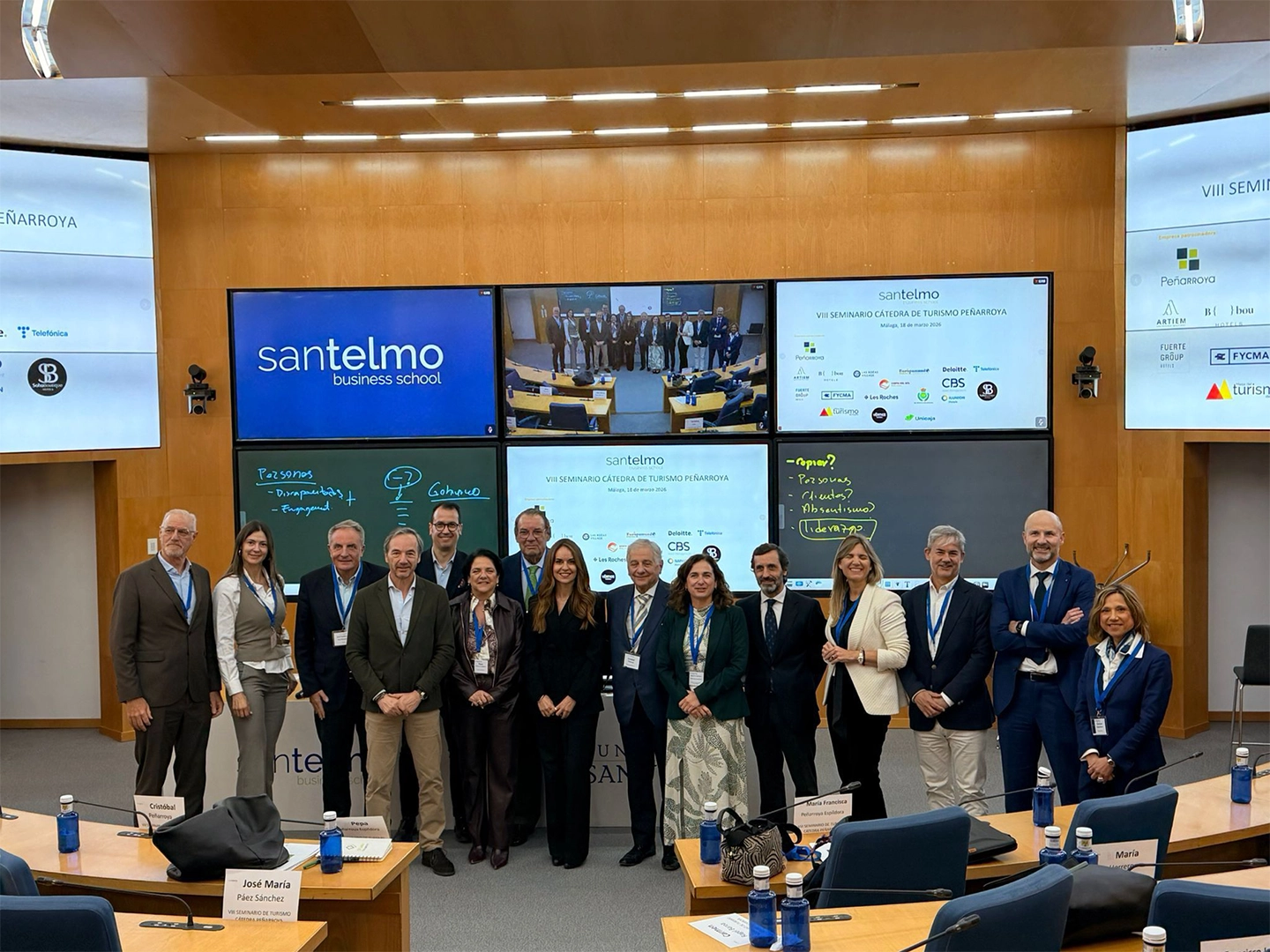 Grupo de personas posando en el centro de un aula tipo auditorio, con varias pantallas al fondo que muestran el nombre “Santelmo Business School”, logotipos de empresas colaboradoras y una imagen grupal. Mesas con tarjetas identificativas y botellas azules rodean la escena.