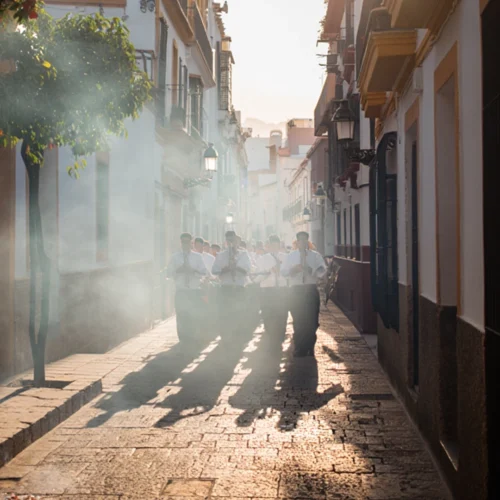 Una banda de música procesional desfila por una calle estrecha y empedrada bajo una intensa luz solar que proyecta largas sombras hacia adelante. Los músicos visten camisas blancas y pantalones oscuros, y tocan instrumentos de viento metal (trompetas) mientras avanzan entre una ligera neblina de incienso o humo. A la izquierda, un naranjo con frutos visibles decora la acera, y los edificios blancos de estilo tradicional flanquean el recorrido, creando una atmósfera solemne y luminosa.