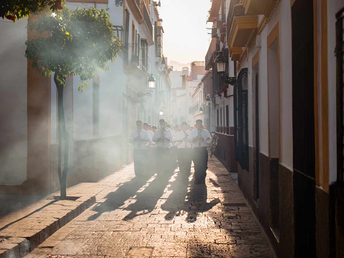 Una banda de música procesional desfila por una calle estrecha y empedrada bajo una intensa luz solar que proyecta largas sombras hacia adelante. Los músicos visten camisas blancas y pantalones oscuros, y tocan instrumentos de viento metal (trompetas) mientras avanzan entre una ligera neblina de incienso o humo. A la izquierda, un naranjo con frutos visibles decora la acera, y los edificios blancos de estilo tradicional flanquean el recorrido, creando una atmósfera solemne y luminosa.
