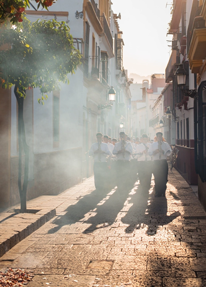Una banda de música procesional desfila por una calle estrecha y empedrada bajo una intensa luz solar que proyecta largas sombras hacia adelante. Los músicos visten camisas blancas y pantalones oscuros, y tocan instrumentos de viento metal (trompetas) mientras avanzan entre una ligera neblina de incienso o humo. A la izquierda, un naranjo con frutos visibles decora la acera, y los edificios blancos de estilo tradicional flanquean el recorrido, creando una atmósfera solemne y luminosa.