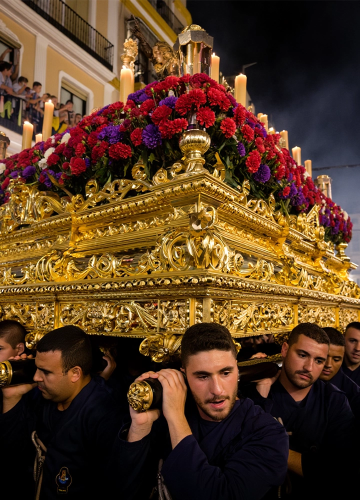 Un grupo de hombres carga sobre sus hombros un pesado paso de Semana Santa tallado en madera dorada y decorado con abundantes flores rojas y moradas. La escena ocurre de noche, con cirios encendidos iluminando la estructura, mientras el público observa desde balcones al fondo.