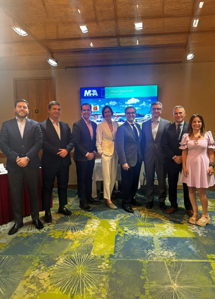 Grupo de personas posando en una sala de conferencias con iluminación cálida. Visten ropa formal y están alineadas frente a una pantalla que muestra una presentación con imágenes de un destino costero y el texto Vision Opportunity. En primer plano, una alfombra colorida con motivos geométricos.