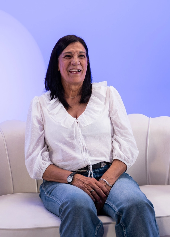 Persona sentada en un sofá claro de diseño curvo, con blusa blanca y vaqueros, las manos entrelazadas sobre el regazo. El fondo es un espacio iluminado en tonos azules suaves que crea un ambiente de estudio sencillo y acogedor.