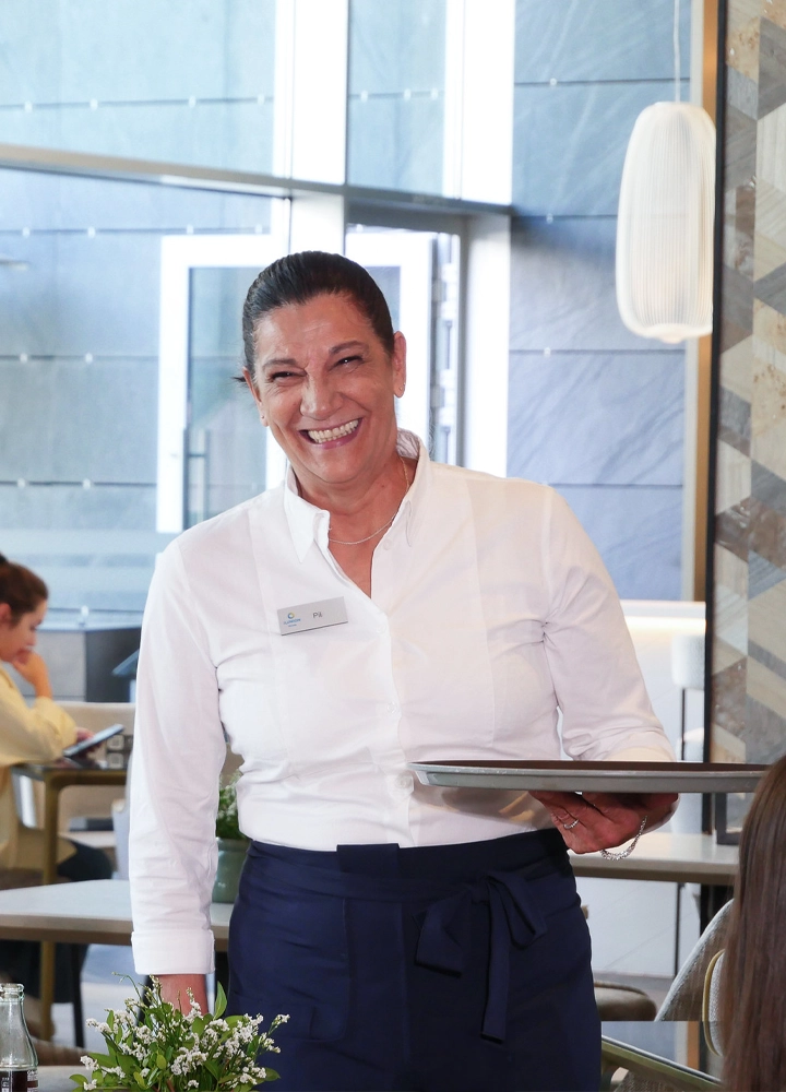 Persona con camisa blanca y delantal azul oscuro sosteniendo una bandeja metálica en un comedor luminoso. El entorno muestra mesas ocupadas, grandes ventanales y una lámpara de diseño en la pared, creando un ambiente moderno y cuidado.