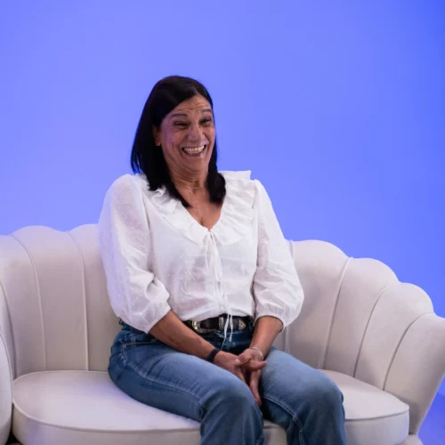 Persona sentada en un sofá claro de diseño curvo, con blusa blanca y vaqueros, las manos entrelazadas sobre el regazo. El fondo es un espacio iluminado en tonos azules suaves que crea un ambiente de estudio sencillo y acogedor.