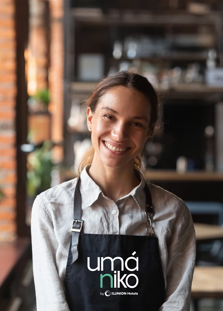 Retrato de primer plano de una mujer joven sonriendo, con una camisa de lino claro y un delantal negro de trabajo. En el delantal se lee el logotipo 