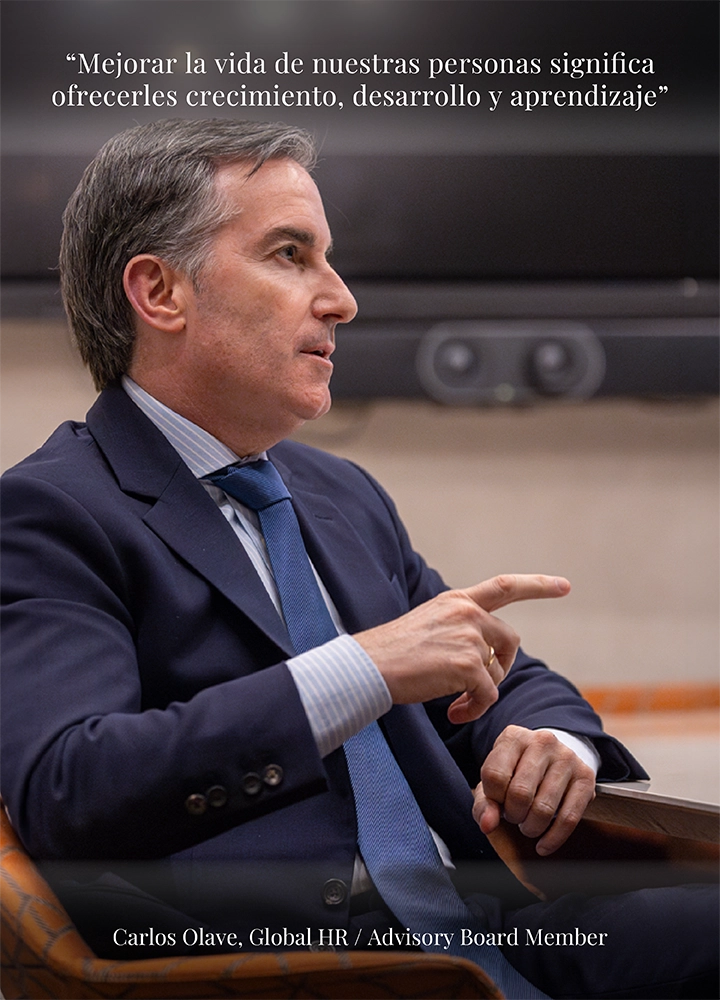 Persona sentada en una sala de reuniones, vestida con traje oscuro y corbata azul, realizando un gesto con la mano mientras participa en una conversación profesional. El entorno es sobrio y corporativo, con una pantalla al fondo y una mesa delante que refuerza el contexto de diálogo estratégico. En la parte superior aparece la cita “Mejorar la vida de nuestras personas significa ofrecerles crecimiento, desarrollo y aprendizaje”, y en la parte inferior el texto “Carlos Olave, Global HR / Advisory Board Member”, transmitiendo un mensaje de liderazgo, compromiso con las personas y desarrollo profesional.
