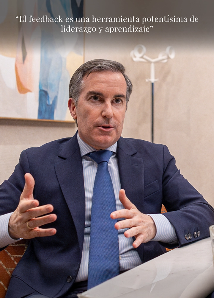 Hombre con traje azul marino y corbata azul claro gesticulando con ambas manos mientras está sentado frente a una mesa clara. A su espalda se ve una pared con un cuadro abstracto en tonos suaves y un perchero blanco de diseño minimalista. En la parte superior aparece la frase: “El feedback es una herramienta potentísima de liderazgo y de aprendizaje”.