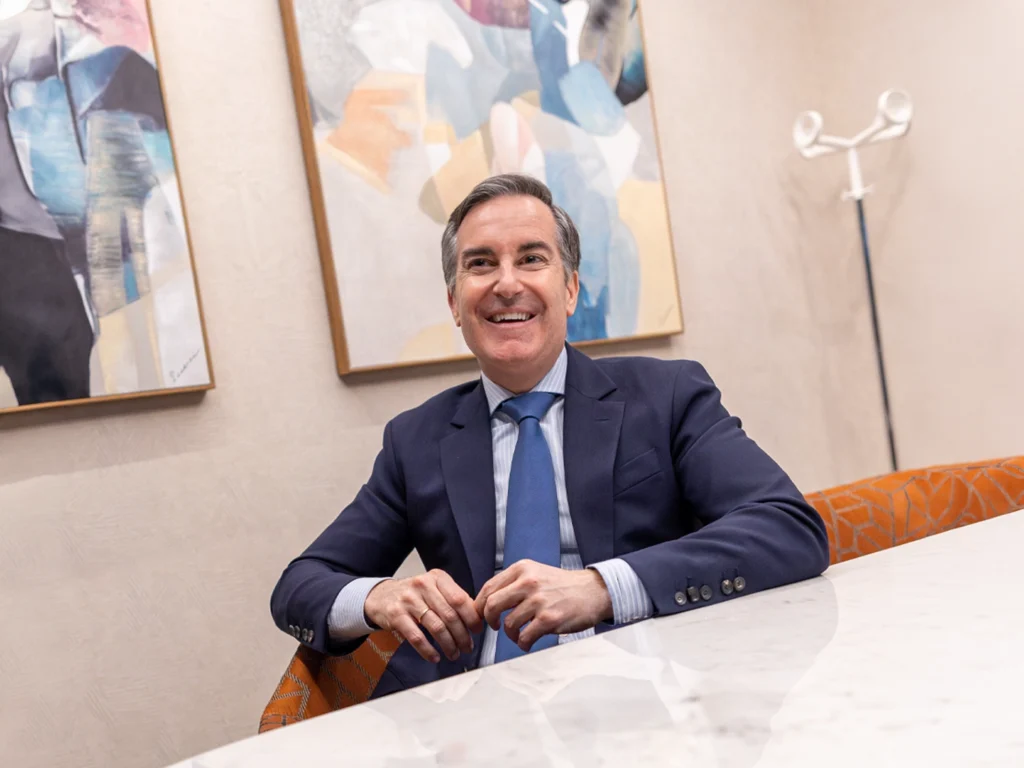 Hombre con traje azul marino y corbata azul claro sentado ante una mesa blanca, con las manos entrelazadas; detrás hay dos cuadros abstractos y un perchero blanco en una sala de estilo moderno.